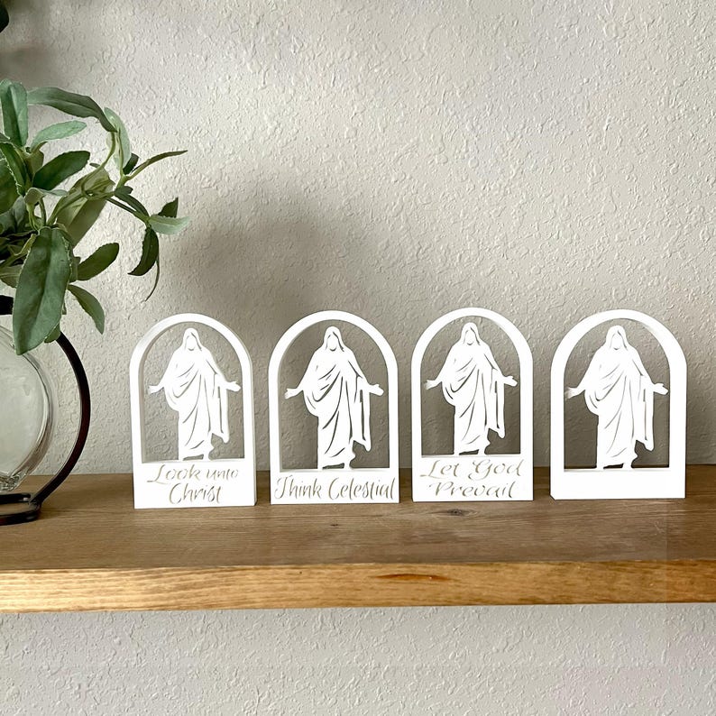 May include: Four white, arched figurines with a religious theme. Each features a figure with outstretched arms and text: "Look unto Christ", "Think Celestial", and "Let God Prevail". They are displayed on a wooden shelf with a plant in the background.