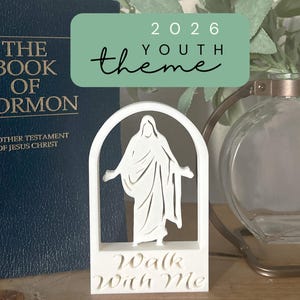 May include: A white, arched figurine depicting a figure with outstretched arms, with the words "Walk With Me" at the base. The background includes a dark blue book and a decorative glass vase. A green banner reads "2026 Youth Theme".