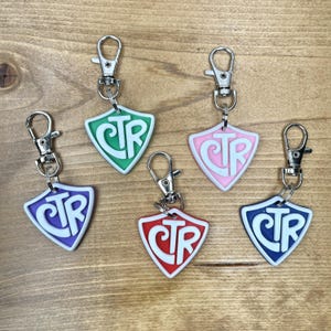 May include: A collection of five shield-shaped keychains in various colors: green, pink, red, purple, and blue. Each keychain features the letters "CTR" in white, with a silver clasp for attachment. The keychains are displayed on a wooden surface.