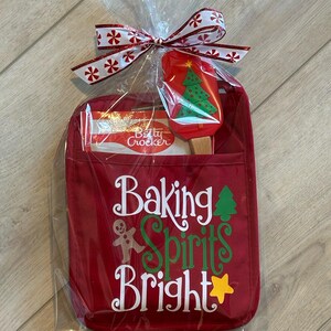 May include: A red oven mitt gift set with a Christmas theme. The mitt has the words "Baking Spirits Bright" and a gingerbread man graphic. A spatula with a Christmas tree design and a Betty Crocker baking mix are included. The set is wrapped in cellophane and tied with a festive ribbon.
