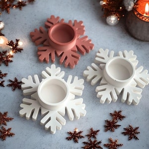 May include: Three snowflake-shaped candle holders in the image. Two are white, and one is a muted rose color. Each holder has a circular well in the center for a candle. Decorative snowflakes are scattered around the holders, suggesting a winter or holiday theme.