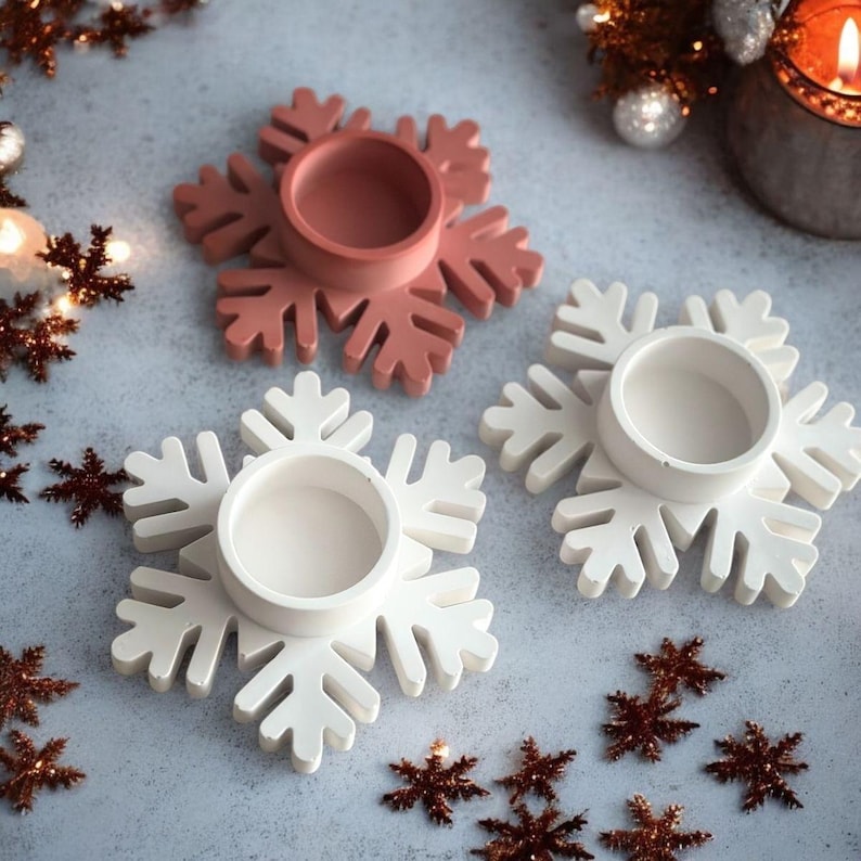 May include: Three snowflake-shaped candle holders in the image. Two are white, and one is a muted rose color. Each holder has a circular well in the center for a candle. Decorative snowflakes are scattered around the holders, suggesting a winter or holiday theme.