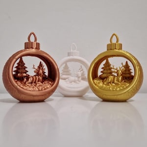 May include: Three Christmas ornaments in bronze, white, and gold. Each ornament features a winter scene with deer and pine trees inside. The ornaments have a loop at the top for hanging. The ornaments are on a white surface.