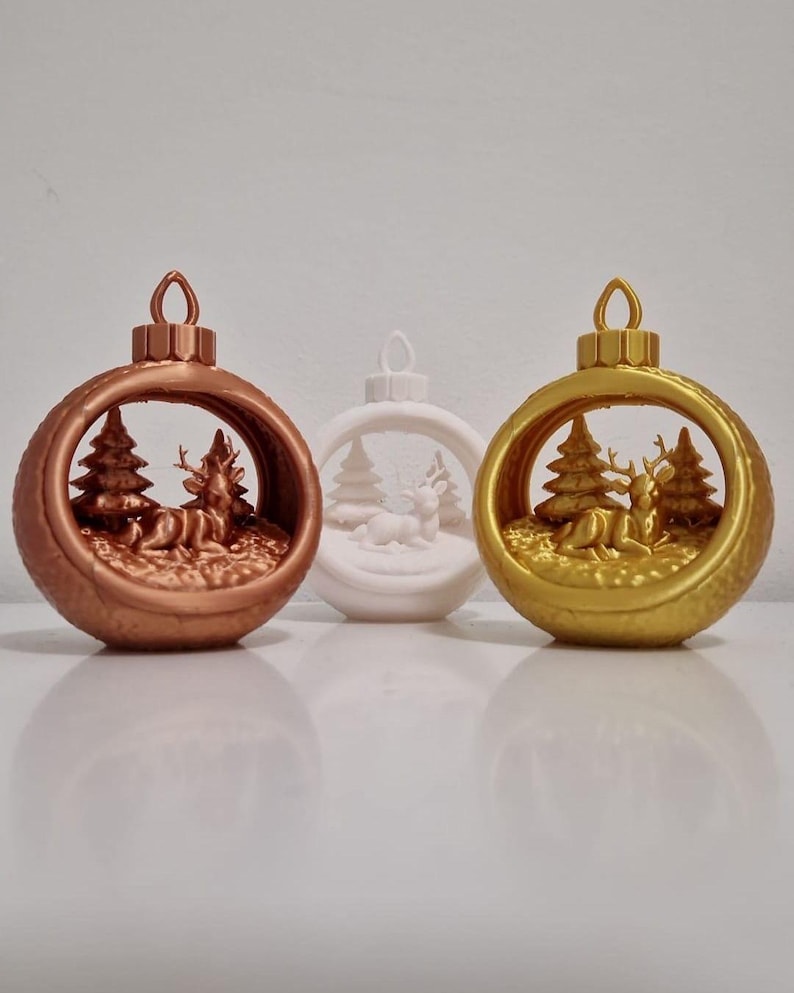 May include: Three Christmas ornaments in bronze, white, and gold. Each ornament features a winter scene with deer and pine trees inside. The ornaments have a loop at the top for hanging. The ornaments are on a white surface.