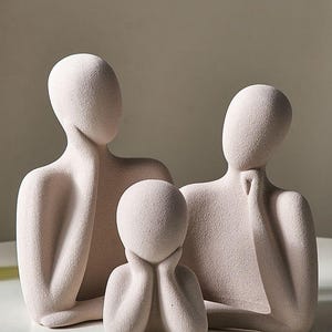 May include: A set of three abstract sculptures in a neutral color. The figures have smooth, rounded forms, with the two larger figures resting their chins on their hands. The smallest figure has its hands under its chin. The sculptures are on a white surface.