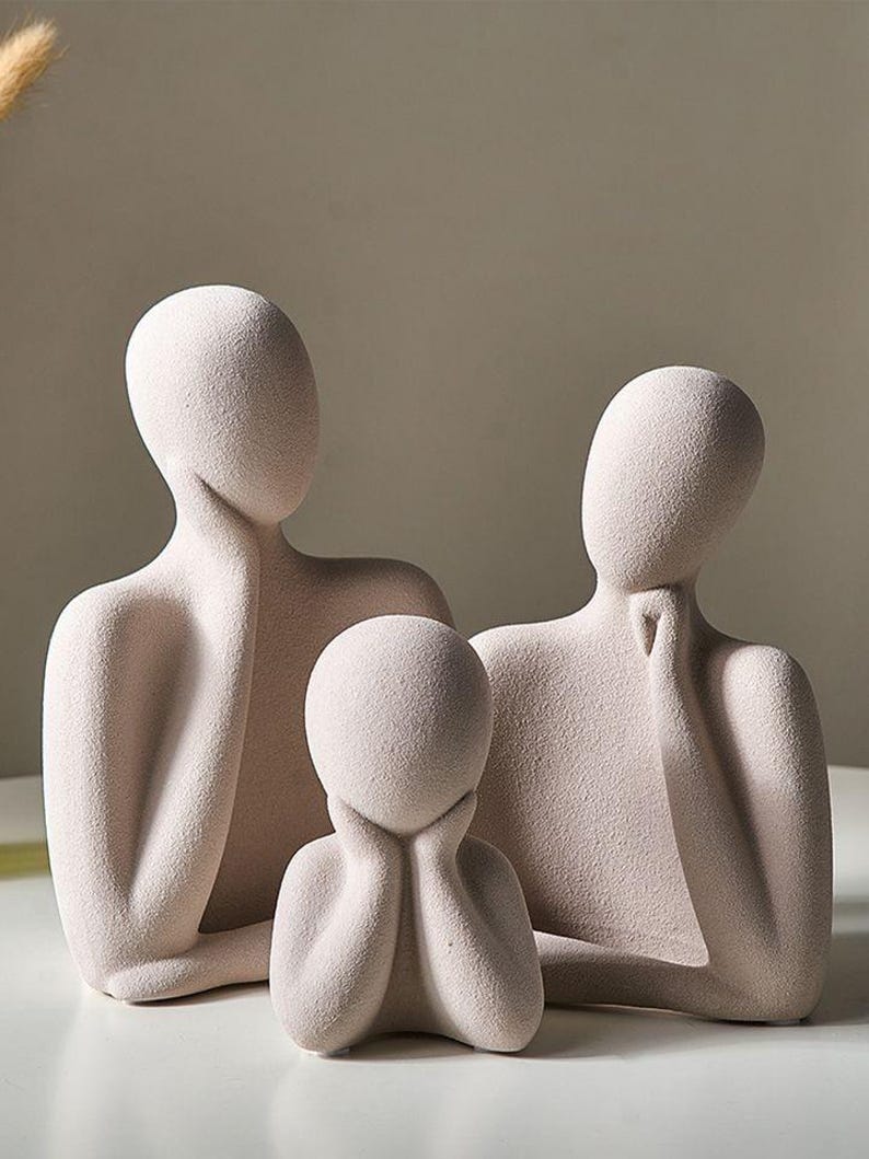 May include: A set of three abstract sculptures in a neutral color. The figures have smooth, rounded forms, with the two larger figures resting their chins on their hands. The smallest figure has its hands under its chin. The sculptures are on a white surface.