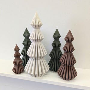 May include: A collection of decorative, geometric Christmas trees in varying sizes and colors. The trees are shades of brown, green, and off-white, with a textured, layered design. They are arranged on a white shelf against a white wall.