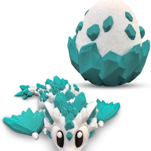 May include: A white and teal 3D-printed dragon toy with large eyes and crystal-like accents. Beside it is a matching egg-shaped container with teal crystal-like accents. The dragon has wings and a long body.