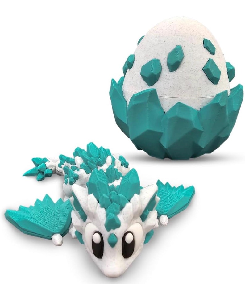 May include: A white and teal 3D-printed dragon toy with large eyes and crystal-like accents. Beside it is a matching egg-shaped container with teal crystal-like accents. The dragon has wings and a long body.