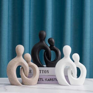 May include: A collection of abstract family sculptures in neutral tones. The sculptures are in black, white, and beige, with a minimalist design. The figures are intertwined, symbolizing unity and connection. The sculptures are displayed on a white surface.