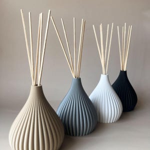 May include: Four decorative reed diffusers in a row, each with a different color vase: beige, gray, white, and black. Each vase has a ribbed design and contains several wooden reeds for fragrance diffusion.