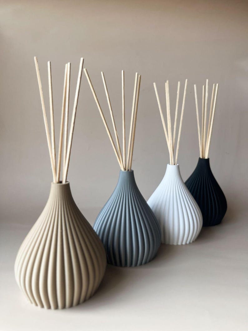 May include: Four decorative reed diffusers in a row, each with a different color vase: beige, gray, white, and black. Each vase has a ribbed design and contains several wooden reeds for fragrance diffusion.