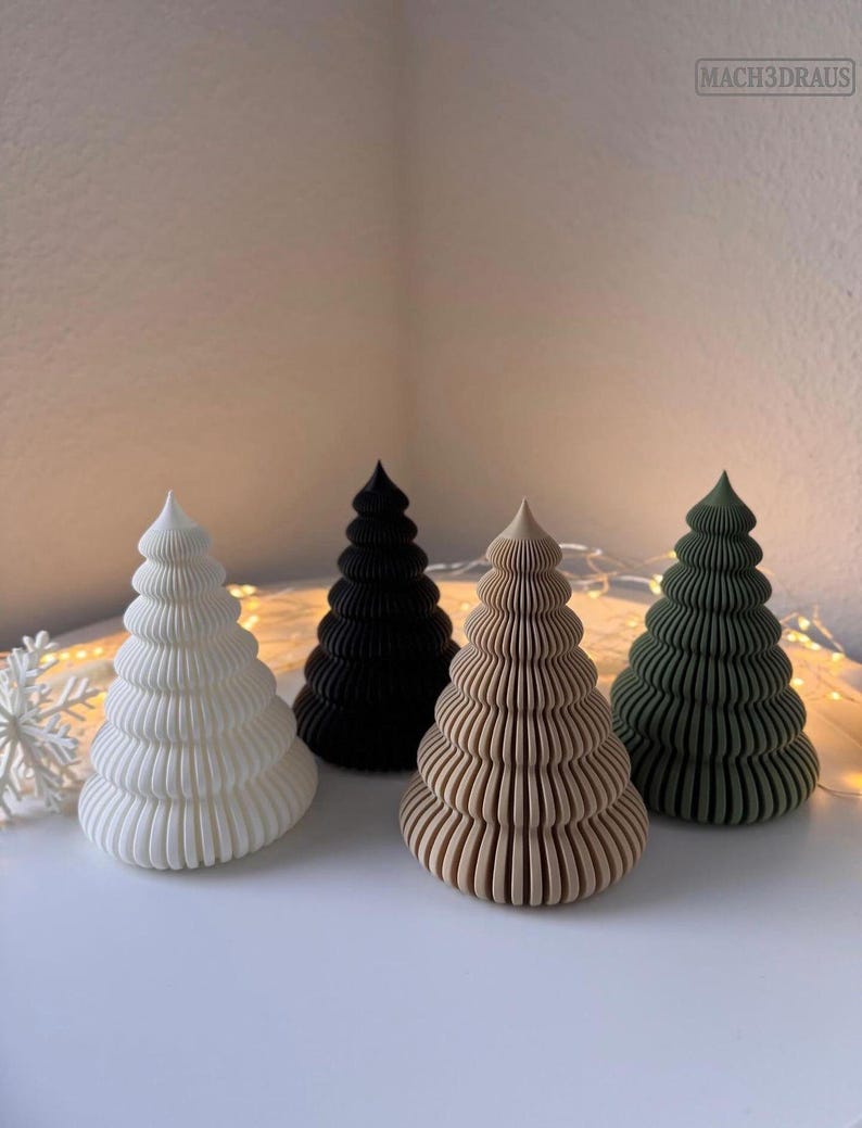 May include: Four decorative Christmas tree figurines in white, black, tan, and green. Each tree has a layered, ridged design. The trees are arranged on a white surface, with a snowflake and string lights in the background. The text "MACH3DRAUS" is in the upper right corner.