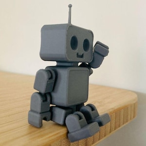 May include: A gray robot figurine with a square head, body, and limbs. The robot has a small antenna and is posed in a sitting position, with its arm raised as if waving. The robot is made of a solid material and sits on a wooden surface.