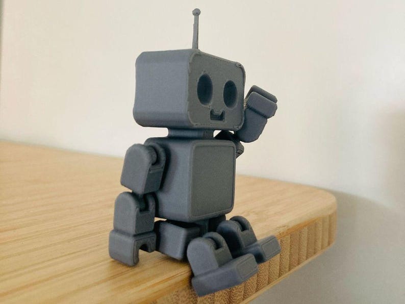 May include: A gray robot figurine with a square head, body, and limbs. The robot has a small antenna and is posed in a sitting position, with its arm raised as if waving. The robot is made of a solid material and sits on a wooden surface.