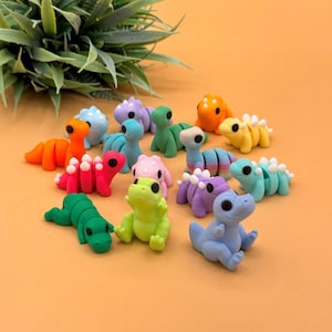 May include: A collection of colorful, miniature dinosaur figurines in various poses. The dinosaurs are in shades of orange, red, pink, green, yellow, blue, and purple. Each dinosaur has a unique design, with some featuring segmented bodies and others with spiky details.