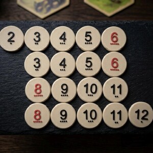 May include: A collection of off-white, circular game pieces with black and red numbers from 2 to 12. Each piece has a number and a series of dots. The pieces are arranged on a dark, textured surface.