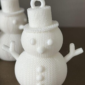 May include: A white snowman ornament with a top hat, arms, and a carrot nose. The snowman has three buttons down the front and a loop at the top for hanging. The ornament has a textured, knitted appearance.