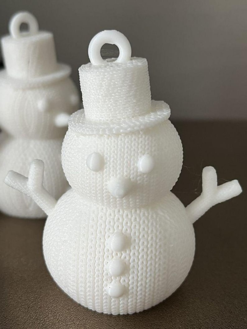 May include: A white snowman ornament with a top hat, arms, and a carrot nose. The snowman has three buttons down the front and a loop at the top for hanging. The ornament has a textured, knitted appearance.