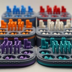 Può includere: Una collezione di colorati pezzi di gioco e organizzatori stampati in 3D. I pezzi sono disposti in vari colori, tra cui blu, arancione, rosso, bianco, viola e verde acqua. Gli organizzatori sono grigi con accenti colorati.