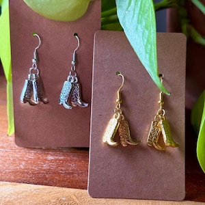 May include: Two pairs of cowboy boot earrings. One pair is silver-coloured, and the other is gold-coloured. The earrings are displayed on brown cardboard earring cards, with green leaves in the background.