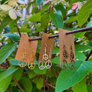 May include: Three pairs of earrings featuring peace sign pendants. The earrings are displayed on brown cardboard tags, suspended from a wooden rod, set against a background of green leaves and pink flowers. The earrings are gold, silver, and bronze.