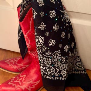 May include: A pair of bright red leather cowboy boots with white decorative stitching on the toe and shaft, draped with a black bandana featuring a white paisley pattern and small scattered blue and green rhinestones throughout the fabric.