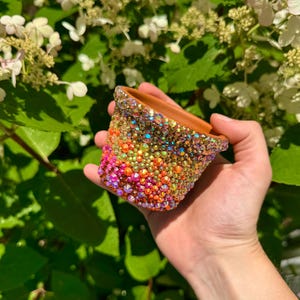 May include: A small, terracotta-colored pot, encrusted with a dazzling array of colorful rhinestones. The rhinestones transition from pink to orange, yellow, and green, creating a vibrant, shimmering effect. The pot is held against a backdrop of green foliage and white flowers.