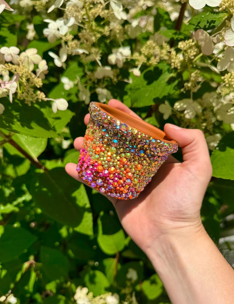 May include: A small terracotta plant pot completely covered in a dense gradient of multi-coloured, iridescent rhinestones. The crystals transition from shades of pink and orange at the base to yellow and clear tones near the top rim.