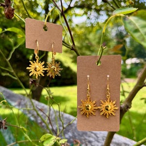 May include: Two pairs of gold-coloured sunburst earrings displayed on brown tags. One pair features hoop closures, while the other has a hook design. The earrings are hanging from a tree branch against a blurred green background.