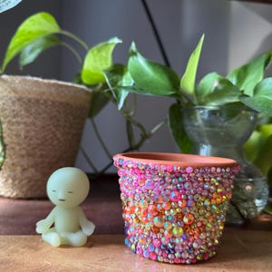 May include: A small terracotta plant pot covered in iridescent, multi-coloured rhinestones in shades of pink, orange, and yellow, positioned next to a small, pale green, smooth-surfaced figurine of a person sitting in a meditative pose.