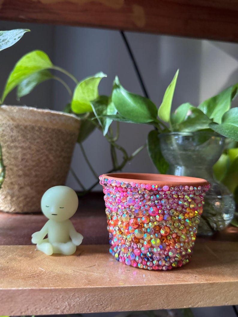 May include: A small terracotta plant pot covered in iridescent, multi-coloured rhinestones in shades of pink, orange, and yellow, positioned next to a small, pale green, smooth-surfaced figurine of a person sitting in a meditative pose.