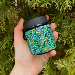 May include: A small jar with a black lid, encrusted with iridescent green and blue rhinestones. The jar is held in a hand against a backdrop of green foliage. The rhinestones create a sparkling, textured surface.
