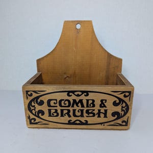 Puede incluir: Soporte de madera para peine y cepillo con acabado natural. La caja rectangular tiene un óvalo negro decorativo con las palabras "COMB & BRUSH" en una fuente serif. El soporte tiene un asa con un agujero para colgar.