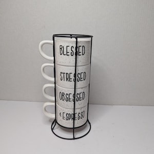 May include: A set of four white espresso cups stacked in a black metal holder. Each cup has a different black text: "BLESSED", "STRESSED", "OBSESSED", and "ESPRESSO". The cups have a speckled design.
