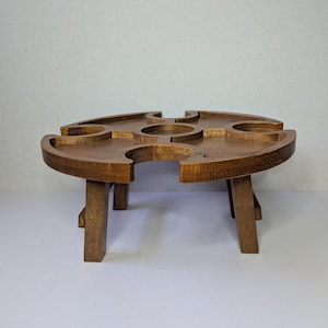 May include: A round, wooden wine table with four curved cutouts for glasses and a central space. The table is supported by four sturdy, rectangular legs. The wood has a warm, brown tone, and the design is simple and functional.