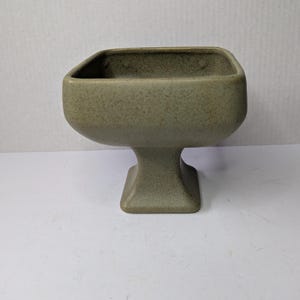 May include: A speckled, olive-green ceramic pedestal bowl. The bowl has a square top with rounded corners and sits on a pedestal base. The base is square and tapers slightly towards the top. The bowl is likely for decorative use.