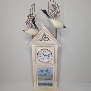 May include: A decorative clock with a cream-colored wooden frame, featuring a round clock face with Roman numerals and a picture of flying ducks. Two metal geese with ornate wings are perched above the clock.