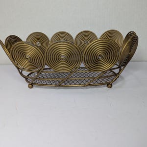 May include: A decorative gold-colored metal basket with a spiral design. The basket has a wire mesh base and is supported by small, rounded feet. The sides feature large, circular spirals, creating a unique and elegant look.