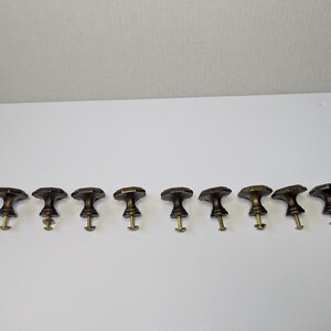 May include: A set of nine dark bronze-toned metal cabinet knobs arranged in a horizontal row. Each knob features a faceted, octagonal top surface and a tapered base, with a threaded brass-colored metal screw extending from the bottom of each piece.