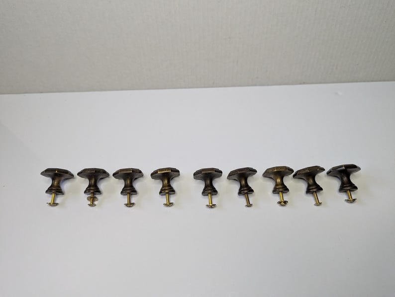 May include: A set of nine dark bronze-toned metal cabinet knobs arranged in a horizontal row. Each knob features a faceted, octagonal top surface and a tapered base, with a threaded brass-colored metal screw extending from the bottom of each piece.
