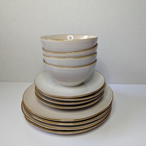 May include: A stack of ceramic dinnerware featuring light gray bowls and plates with a subtle horizontal ribbed texture and a thin, dark brown rim on each piece. The set includes multiple bowls stacked on top of several larger dinner plates.