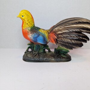 May include: A colorful ceramic rooster figurine. The rooster has a yellow head, orange chest, blue wings, and a long, detailed tail. It is perched on a dark green base with leaf details. The figurine is a decorative item.