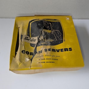 May include: A vintage yellow box of "Corn Servers" with a black and white illustration of a person serving food. The box is torn and shows the text "complete service for four", including corn husk dishes and skewers.