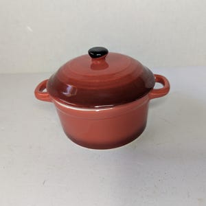 May include: A small, round, red ceramic pot with a matching lid. The lid has a black knob and the pot has two small handles. The pot is designed for cooking and serving food.