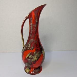 Może przedstawiać: Wysoki, ceramiczny dzban z żywym czerwonym i brązowym szkliwem. Dzbanek ma zakrzywiony uchwyt i rozszerzający się dzióbek. Podstawa ma kształt cokołu. Ogólny design jest w stylu modernizmu z połowy wieku.