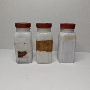 May include: Three vintage white glass jars with red lids. The jars have a rectangular shape and are slightly aged, with some discoloration and labels. The jars are arranged side by side on a white surface.