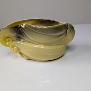 May include: A pale yellow ceramic planter with a smooth, glossy finish and a dark grey airbrushed gradient along the upper rim. The vessel features a sculptural, asymmetrical design with prominent swirling, wave-like embossed details.