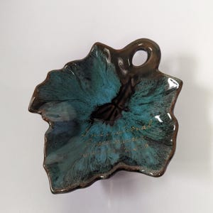 May include: A leaf-shaped ceramic dish with a glossy glaze featuring a dark brown border and a vibrant teal center. The dish has a small handle with a hole and includes the gold cursive text International Peace Garden on the surface.