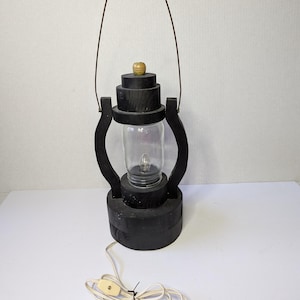 May include: A black wooden lantern with a clear glass jar in the center, housing a lightbulb. The lantern has a metal handle and a wooden base. A white electrical cord with a beige plug extends from the base.